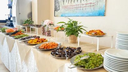 Buffet con varie verdure cotte e piatti impilati su tavolo con tovaglia.