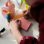 Bambina che scrive su foglietto rosa con giocattoli e casa di plastica sul tavolo