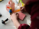 Bambina che scrive su foglietto rosa con giocattoli e casa di plastica sul tavolo